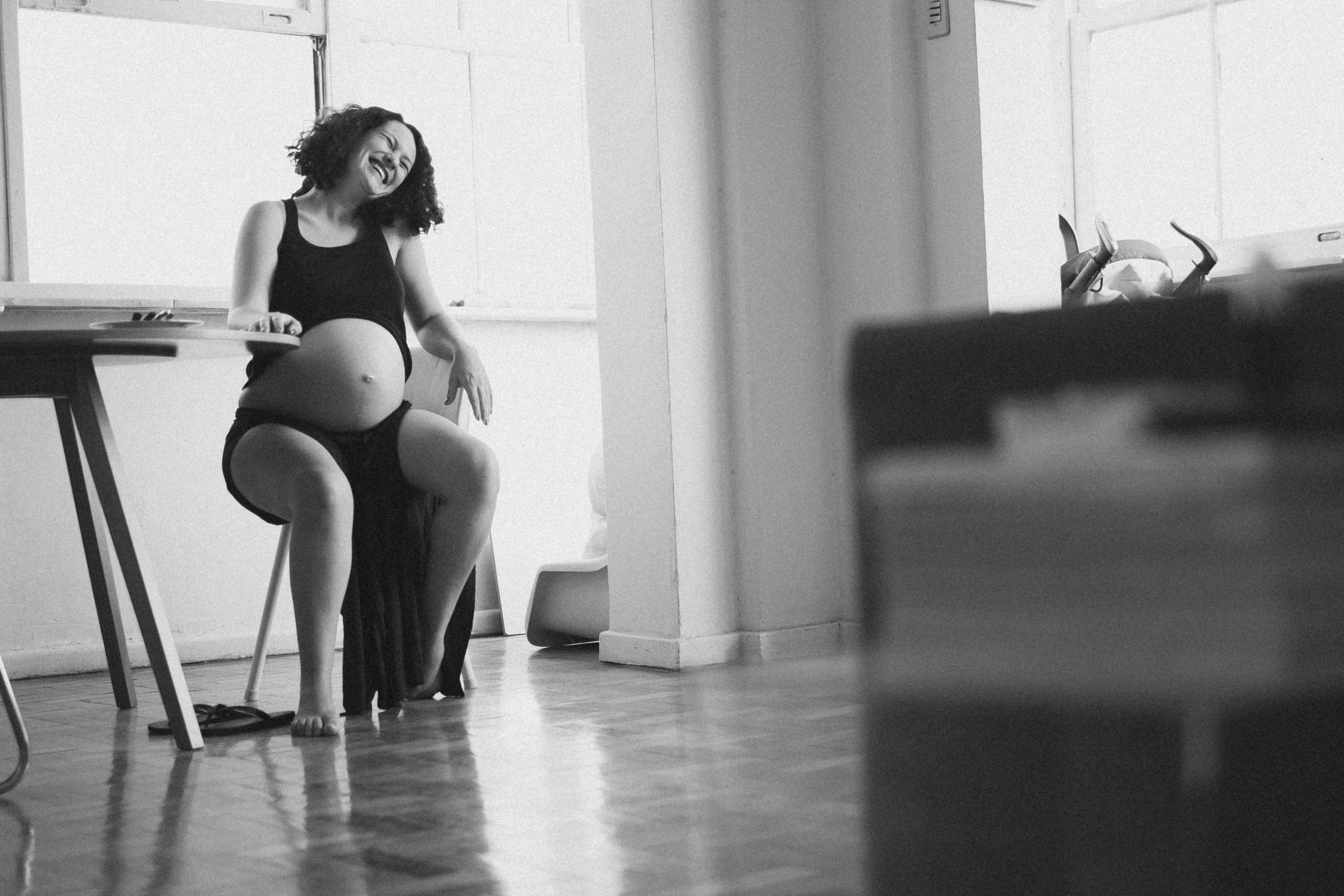 Fotografia de uma mulher grávida de cabelos cacheados sentada em uma cadeira em um apartamento minimalista. Ela usa regata preta e shorts e dá uma gargalhada. Uma mesa de madeira simples está à sua esquerda. Ao fundo, uma janela iluminada e parte de uma parede branca completam a cena.