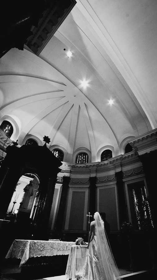 Noiva diante de um altar em uma igreja grandiosa, vista de um ângulo baixo. O altar com toalha rendada domina o primeiro plano, enquanto o fundo exibe um teto abobadado detalhado e janelas arqueadas. A luz suave, vinda do teto, destaca a figura da noiva contra o altar escuro, o noivo ao seu lado. Molduras clássicas adornam as paredes, e há um pequeno crucifixo no altar.