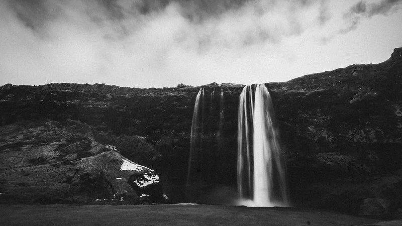 Foto de uma cachoeira que desce por um penhasco escuro. No fundo, penhascos texturizados dominam a cena sob um céu carregado de nuvens. A cachoeira, com múltiplos fluxos, contrasta com as rochas escuras e o céu nublado. Há uma vegetação discreta nas laterais do penhasco.
