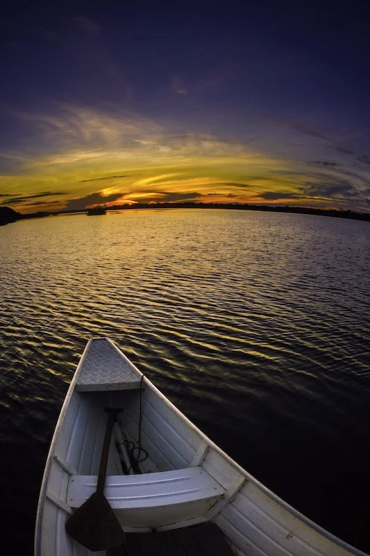 Foto de uma canoa branca de alumínio sobre águas calmas. O pôr do sol ilumina o céu com tons dourados e azuis escuros. Ao fundo, silhuetas de árvores margeiam a água. Ondas suaves refletem as cores vibrantes do céu.