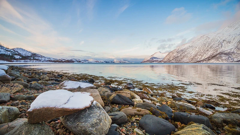 Foto de uma paisagem tranquila em uma costa da Noruega. Rochas lisas em tons de cinza, algumas com neve, dominam o primeiro plano. Ao fundo, um corpo d'água reflete o céu claro e montanhas cobertas de neve. Montanhas mais distantes surgem com picos brancos. As cores predominantes são azuis e cinzas no céu e na água, contrastando com os marrons e cinzas das rochas.