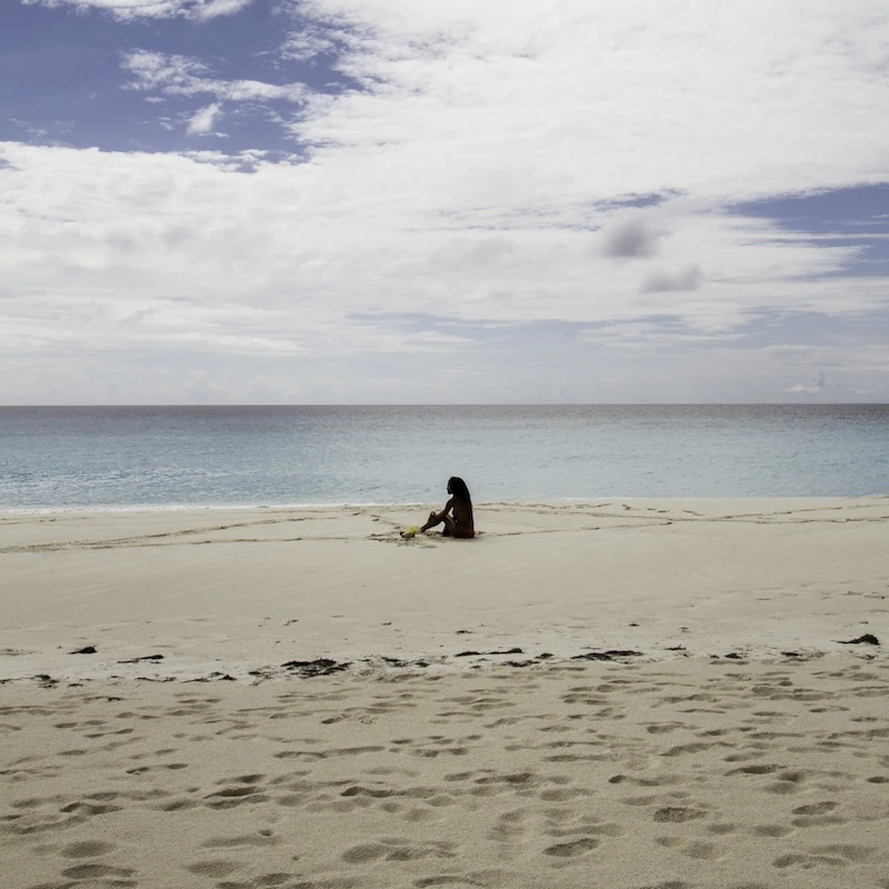 Fotografia de uma praia com mar azulado, e uma silhueta de mulher sentada no meio do quadro. Cores reais.