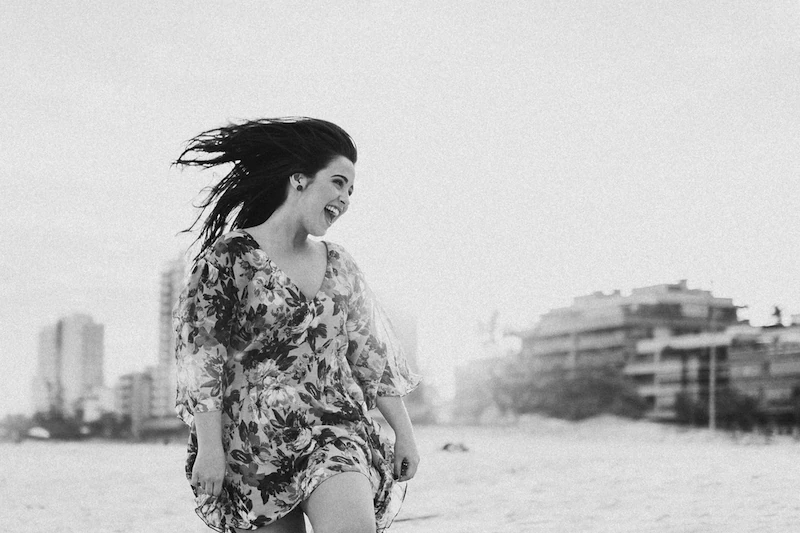 Foto de uma mulher usando um vestido leve florido, esvoaçante. Ela sorri olhando para o lado enquanto caminha em direção à câmera.