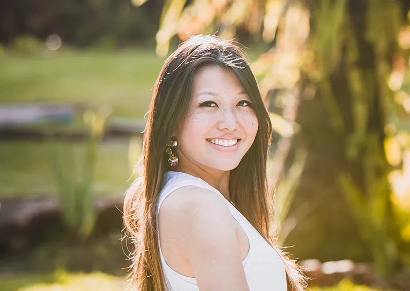 Retrato de uma mulher olhando para a câmera e sorrindo, seus cabelos lisos estão com um brilho dourado da luz do sol que vem de trás.