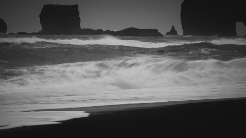 Praia com areias pretas, várias rochas de formatos diversos ao fundo e ondas enevoadas batendo.