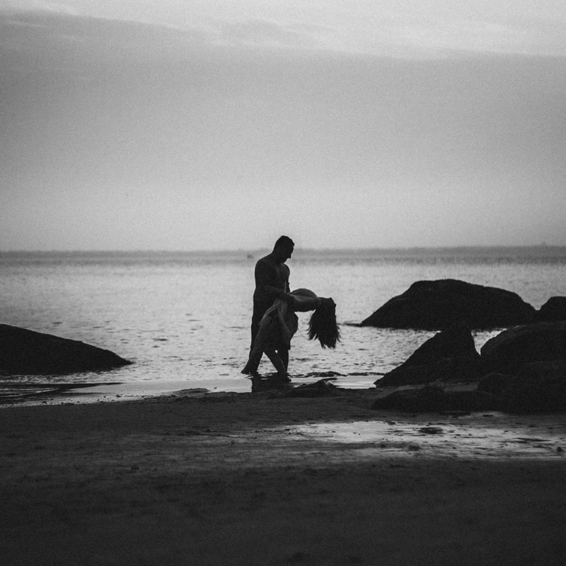 Foto na praia de um casal dançando, onde se vê o mar, pedras e a silhueta do casal.