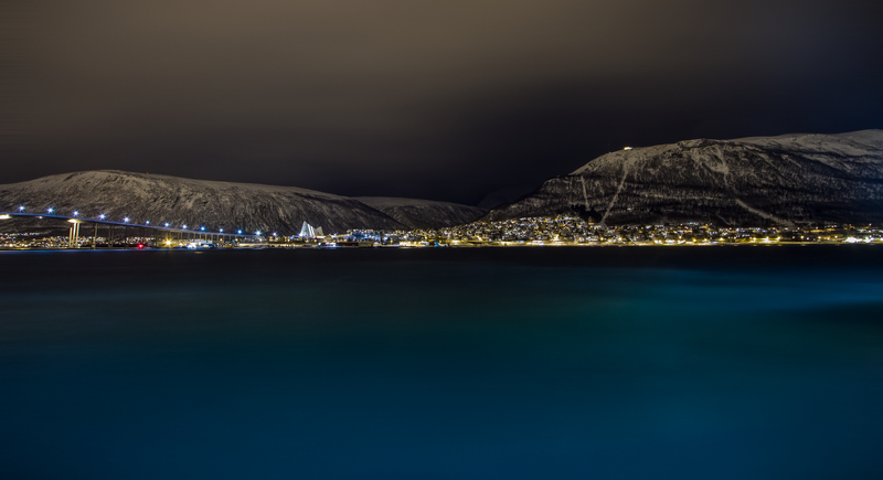 Fotografia noturna de uma cidade vista através do estreito de Tromsøysundet. No primeiro plano, as águas escuras e tranquilas refletem as luzes da cidade, criando uma atmosfera serena. Ao fundo, uma cadeia de montanhas cobertas de neve sob um céu escuro, com Tromsø iluminada à sua base, suas luzes cintilando como estrelas. Uma longa ponte cruza a água, conectando a cidade ao outro lado.