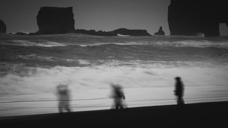 Foto de uma praia, em preto e branco, em que se vê o mar bastante agitado, grandes pedras ao fundo e três crianças encapuzadas correndo.