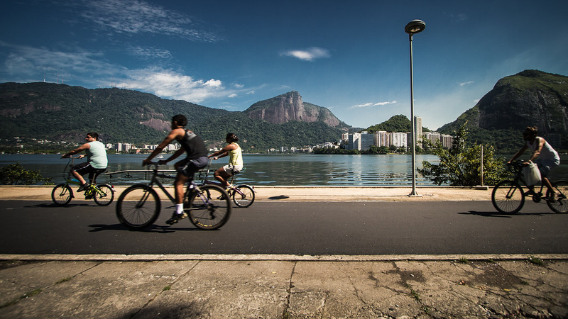 Foto em que se vê a lagoa Rodrigo de Freitas, montanhas ao fundo e uma ciclovia no plano frontal, com ciclistas passando.