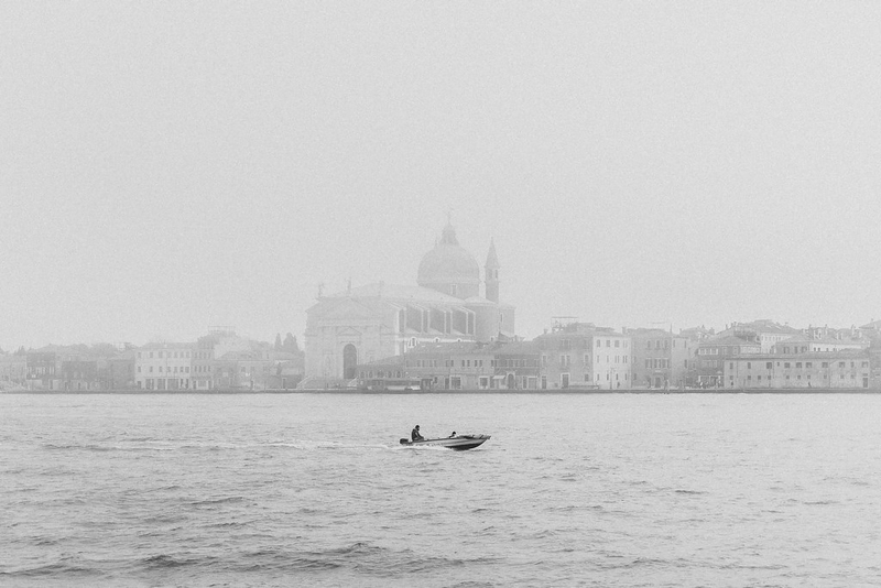 Fotografia serena de um skyline em um dia nublado. No primeiro plano, uma pequena lancha com uma única pessoa desliza pelas águas calmas. Ao fundo, o contorno enevoado do Canal Giudecca, com prédios e a icônica Basílica de Santa Maria della Salute ao longe. A lancha está centralizada e em destaque na foto, mesmo que ocupe pouco espaço na composição.