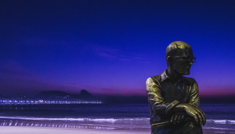 Foto do amanhecer com a praia ao fundo e a estátua de Drummond em primeiro plano.