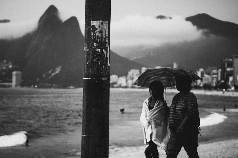 Foto da praia de Ipanema cheia de nuvens e duas moças andando com um guarda-chuva.