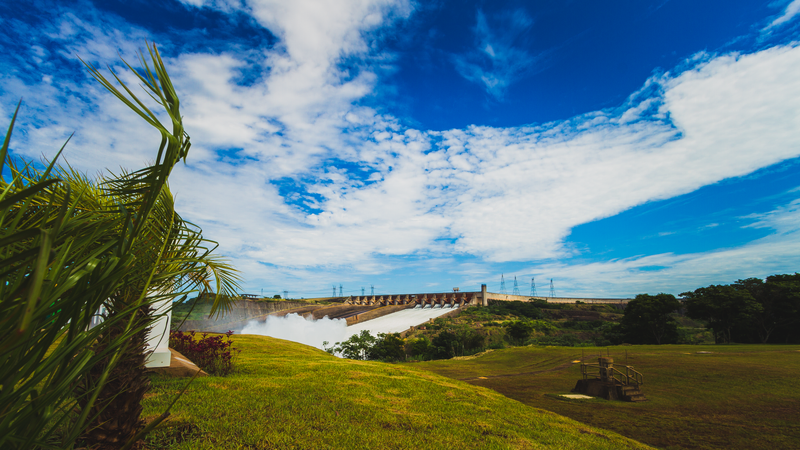 Foto de um vertedouro de hidrelétrica com água jorrando. O céu está azul e, à frente, estão campos esverdeados.