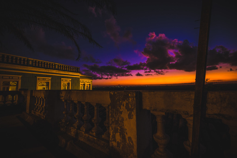 Foto de um pôr do sol onde o céu está indo de alaranjado para rosa para azul, com nuvens brilhando em rosa. Em primeiro plano, um casarão.