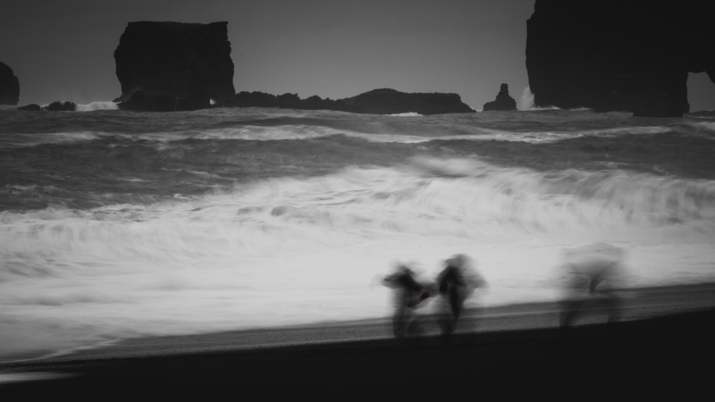 Foto de uma praia com as ondas embaçadas e três figuras humanas também embaçadas.