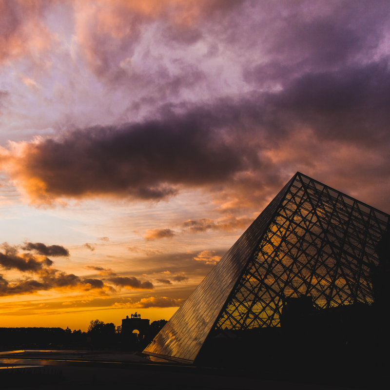A pirâmide de vidro do Louvre iluminada por um céu com nuvens e cheio de cores: laranja, azul, roxo.