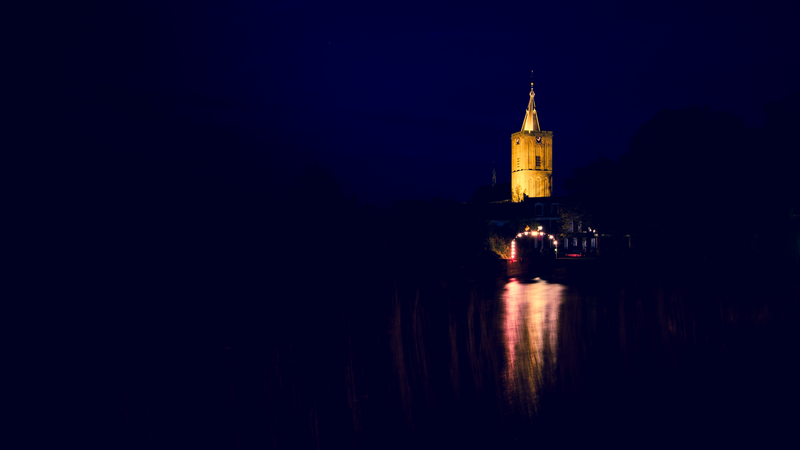 Fotografia de uma torre iluminada contra o céu escuro da noite. No primeiro plano, um canal de água escura reflete o brilho quente da torre. Ao fundo, silhuetas de árvores e prédios completam a cena.