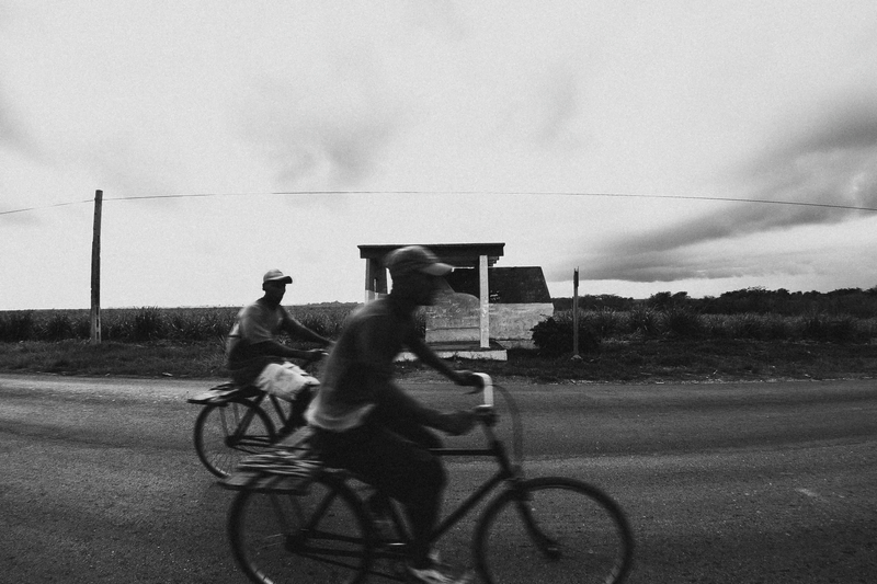 Foto de uma estrada em que se vê uma parada de ônibus vazia. Na frente do quadro passam dois rapazes de boné andando de bicicleta, com o tremor da foto indicando o movimento deles.
