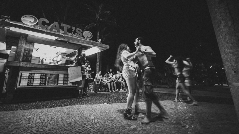 A foto mostra, ao fundo, um quioque de praia com várias pessoas sentadas observando. No plano frontal se vê dois casais dançando agarradinhos. Um deles está à direita, e se vê bem o movimento. O outro está quase no centro do quadro e dá pra ver o movimento nos pés deles.
