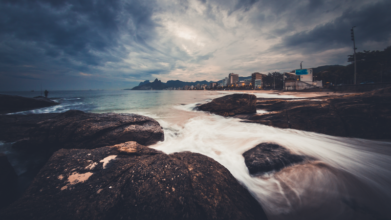 Foto de praia com pedras no primeiro plano, vista da cidade e de montanhas ao fundo.