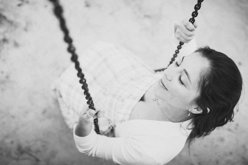 Retrato em preto e branco de uma mulher sorrindo delicadamente. A foto é feita de um ângulo de cima e ela está em um balanço, segurando as correntes.