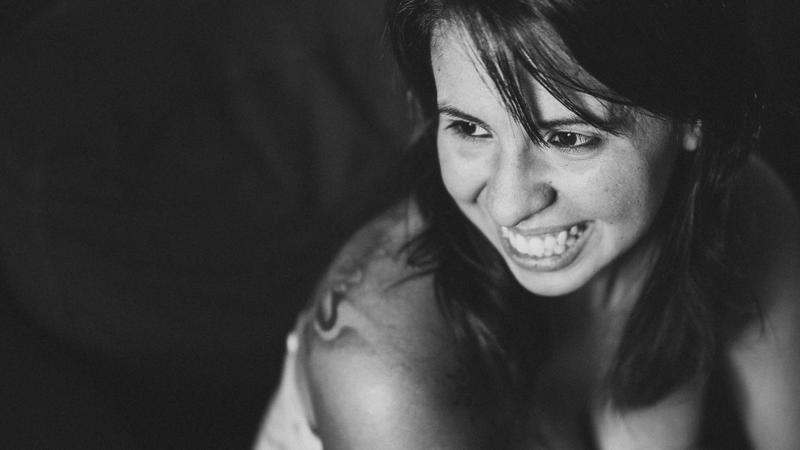 Retrato de mulher de cabelos lisos e franja. Ela dá um grande sorriso e olha para o lado esquerdo do quadro. Somente os olhos estão em foco mas é possível ver uma tatuagem no seu ombro direito.