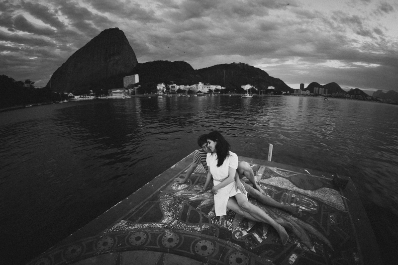 Um homem e uma mulher se abraçam, de olhos fechados, na parte inferior da fotografia. Vemos eles de corpo inteiro e estão sentados no topo de um barco, que tem grafismos pintados. Ao fundo se vê a imensidão da baía de guanabara e o pão de açúcar.