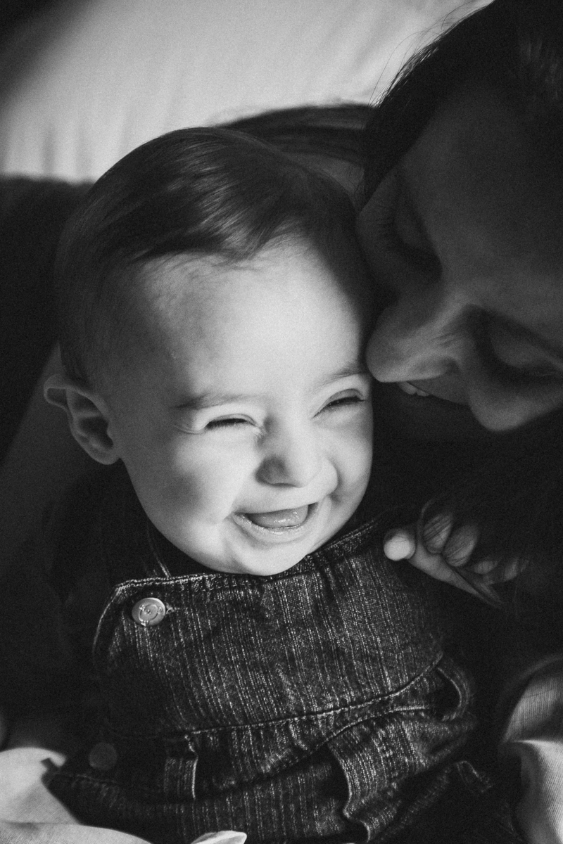 Foto de um bebê sorrindo com a mãe abraçando ele por trás e também sorrindo.