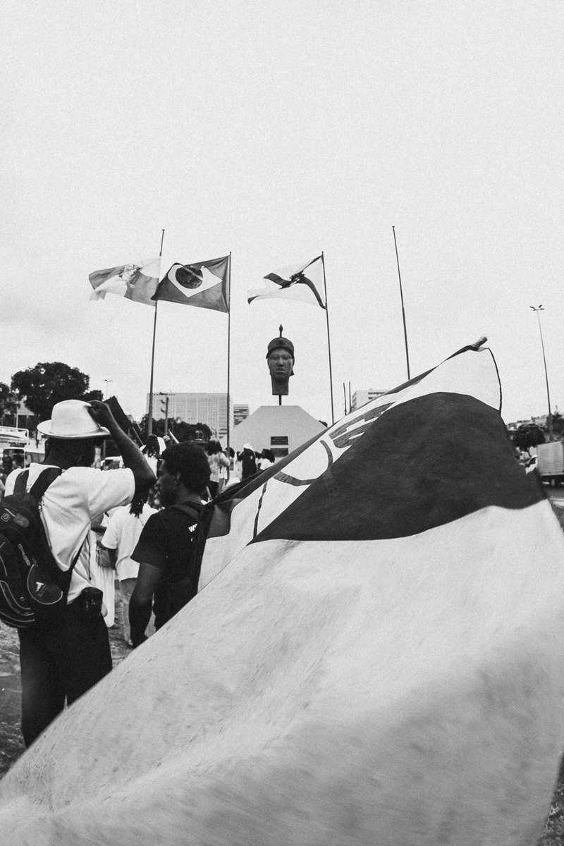 Foto com diversas camadas: na primeira, uma grande bandeira da ONG educafro, na segunda, as pessoas participando da manifestação, na terceira, ao fundo, a estátua de Zumbi e bandeiras do Brasil e do RJ.
