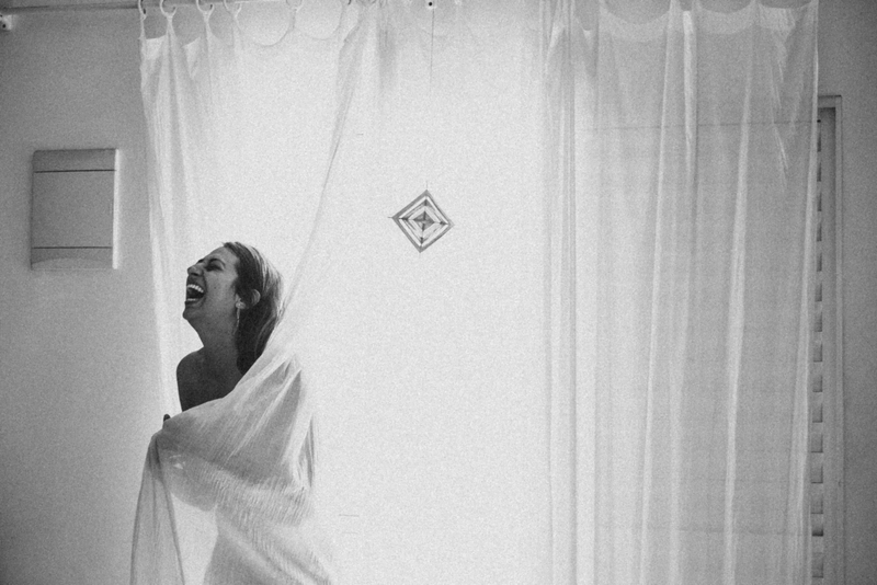 Foto de uma mulher enrolada em uma cortina, a luz vindo de trás, pela janela, e ela dando uma gargalhada.