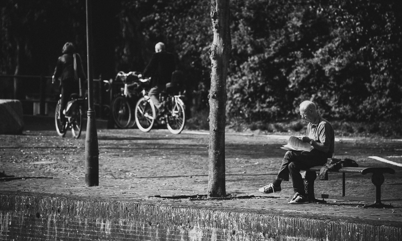 Foto PB de um senhor lendo papéis em um banco e algumas pessoas passando de bicicleta ao fundo.