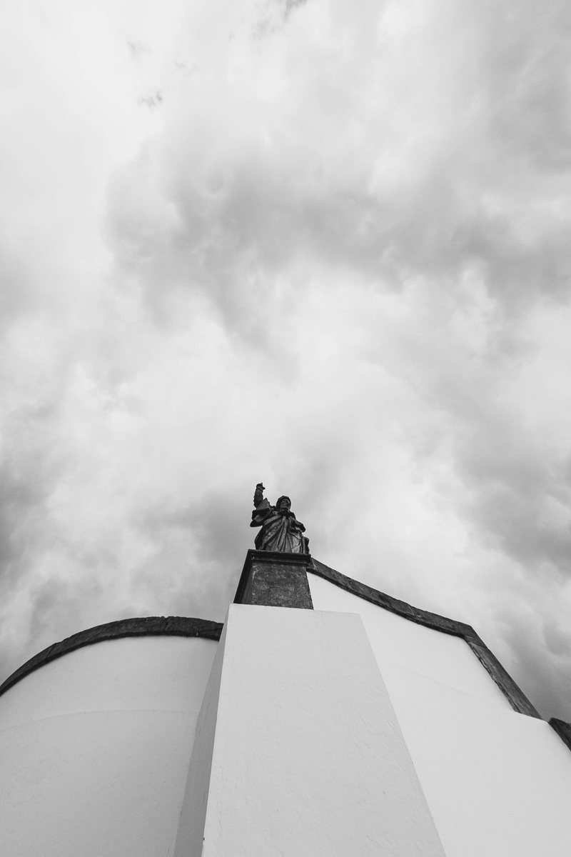 Foto de baixo para cima de uma estátua com o braço levantado, no topo de um muro. A foto tem tons predominantemente claros.