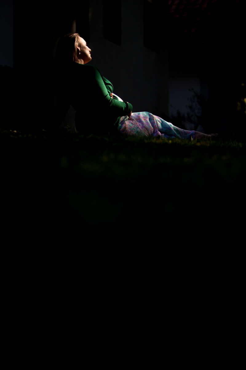 Retrato de uma pessoa grávida deitada na grama, de lado. Ela está olhando para frente, sendo possível ver apenas a silhueta do seu rosto. Ela usa um casaco verde escuro e um vestido florido. A luz é dramática e bastante constrastada, iluminando apenas lateralmente o corpo da modelo e a grama, pouco se vendo do fundo ou da parte frontal.