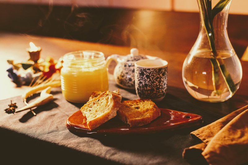 Imagem de uma mesa posta com pães, um pequeno bule e um copo de onde sai fumacinha. É possível ver a base de um vaso transparente com o caule de flores.