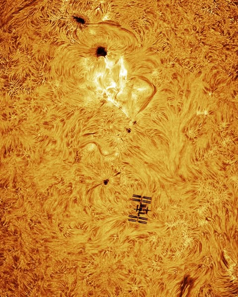 Imagem aproximada do sol com seus flares em destaque e a silhueta da estação espacial, pequenina, no canto inferior direito.