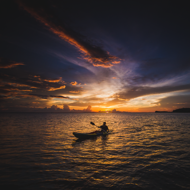 Fotografia mostrando um rio com o pôr do sol alaranjado ao fundo. Em primeiro plano, uma pessoa rema em um caiaque.